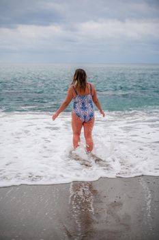 A plump woman in a bathing suit enters the water during the surf. Alone on the beach, Gray sky in the clouds, swimming in winter