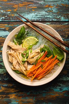 Asian soup with rice noodles, chicken and vegetables in ceramic bowl served with spoon and chopsticks on rustic wooden background from above, Chinese or Thai cuisine