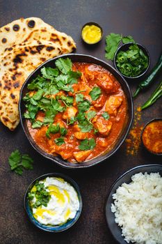 Traditional Indian dish Chicken tikka masala with spicy curry meat in bowl, basmati rice, bread naan, yoghurt raita sauce on rustic dark background, top view, close up. Indian style dinner from above