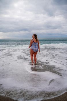 A plump woman in a bathing suit enters the water during the surf. Alone on the beach, Gray sky in the clouds, swimming in winter