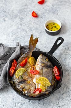 Roasted or grilled fish sea bream or dorado with lemon, herbs and tomatoes in cast iron skillet great for healthy meal or Mediterranean diet on rustic white stone table, angle view
