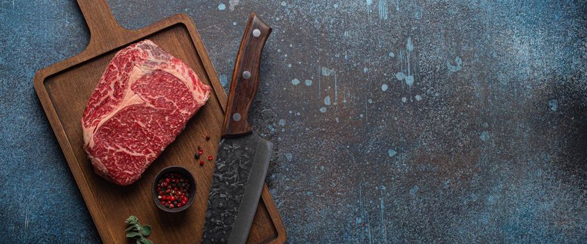 Raw meat beef marbled prime cut steak Ribeye on wooden cutting board and rustic concrete kitchen table background from above with big knife and spices, beefsteak concept