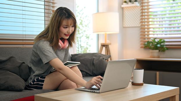Smiling young lady in casual clothes using laptop on couch, spending time on weekend at home.