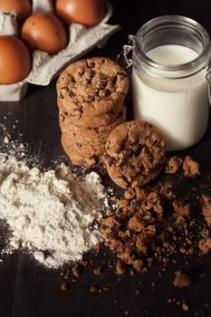 Homemade chocolate chip cookies with bottle of milk, white flour and crumbs on rustic wooden table.