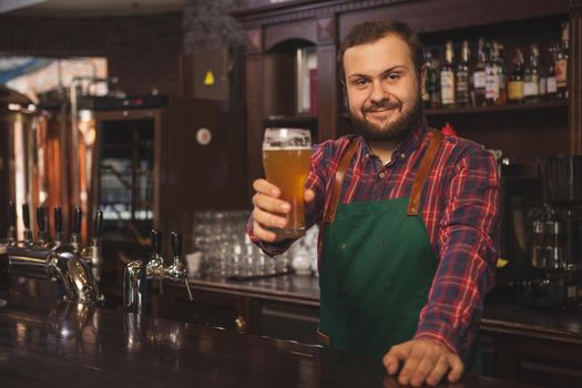 Happy bearded bartender wearing apron, smiling cheerfully to the camera, holding glass of beer, posing behind the counter. Friendly brewer working at his pub, copy space. Recreation concept