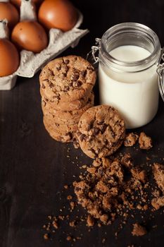 Homemade chocolate chip cookies with bottle of milk and crumbs on rustic wooden table. Fresh eggs.