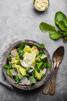 Green organic vegetables salad with avocado, kale, green peas, sprouting herbs, radish in rustic bowl on gray concrete background, close-up, top view. Healthy clean eating, diet or detox concept