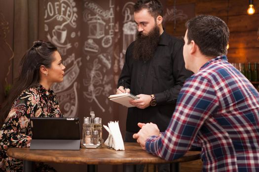 Bearded young waiter taking the order from young couple in restaurant. Vintage pub.