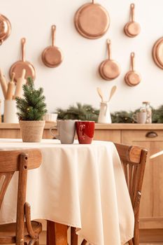 Two tea cups on Christmas table with little tree in bright modern decorated kitchen interior with many copper pans on wall, kitchen tools and utensils, candles, space for text