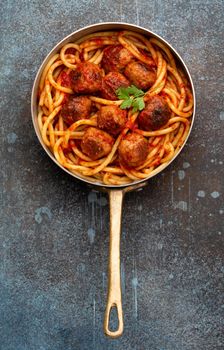 Overhead of traditional homemade pasta with roasted meatballs, tomato sauce, green herbs in copper vintage pan on rustic concrete background. Classic homemade meatballs spaghetti for dinner
