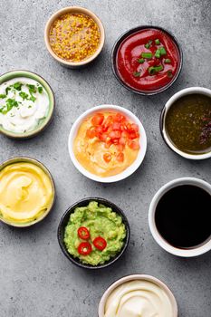 Set of different sauces in bowls on gray rustic concrete background, top view, close-up. Tomato ketchup, mayonnaise, guacamole, mustard, soy sauce, pesto, cheese sauce - assortment of dips