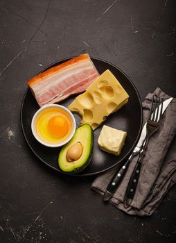 Main keto foods as butter, olive oil, fried egg, avocado, cheese, fat meat bacon for ketogenic diet on black plate on dark rustic stone background, products for healthy weight loss top view