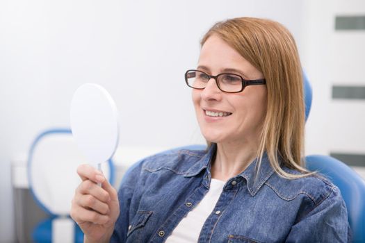 Mature woman looking at her teeth in the mirror at dental clinic