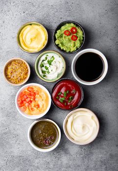 Set of different sauces in bowls on gray rustic concrete background, top view, close-up. Tomato ketchup, mayonnaise, guacamole, mustard, soy sauce, pesto, cheese sauce - assortment of dips