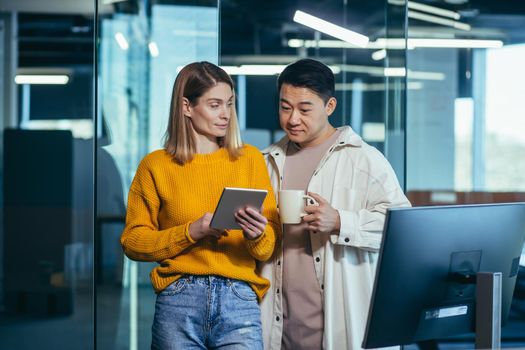 Two employees are having fun. during the break an Asian man and a blonde woman, colleagues work together in a modern office, look at a tablet computer together