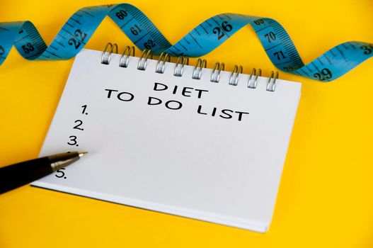 Diet to do list text on white notepad with blue measuring tape on yellow background. Health and diet concept.