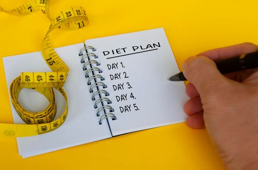 Diet plan text on white notepad with measuring tape on yellow background. Health and diet concept.