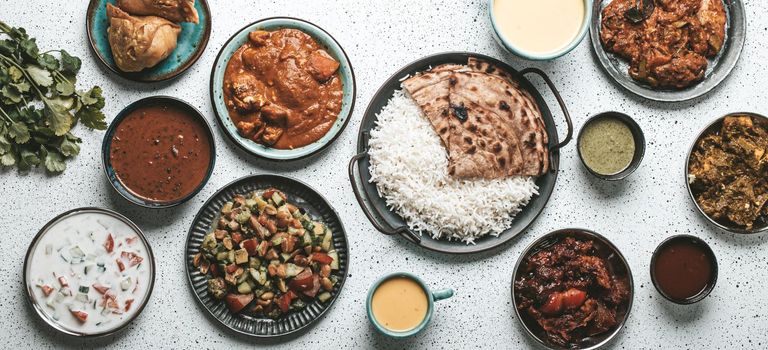 Indian ethnic food buffet on white concrete table from above: curry, samosa, rice biryani, dal, paneer, chapatti, naan, chicken tikka masala, mango lassi, dishes of India for dinner background