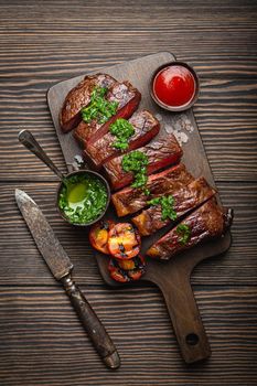 Grilled or fried and sliced marbled meat steak with fork, tomatoes as a side dish and different sauces on wooden cutting board, top view, close-up, wooden rustic background. Beef meat steak concept