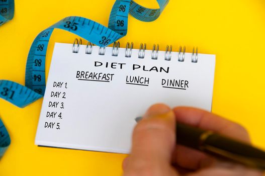 Diet plan on white notepad with blue measuring tape on yellow background. Health and diet concept.
