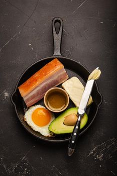 Main keto foods as butter, olive oil, fried egg, avocado, fat meat bacon for ketogenic diet on black cast iron pan on dark rustic stone background, products for healthy weight loss top view