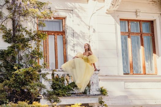 a beautiful smiling and kind woman in a gorgeous yellow dress stands on the balcony of an old vintage house.