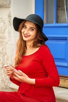 Portrait of tourist young caucasian woman in a red long dress and black hat with suitcase sitting on the stairs outdoors