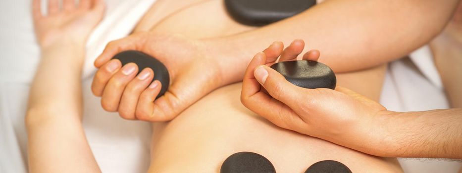 Hot stone massage on the female back with hands of masseur holding black massage stones in spa salon