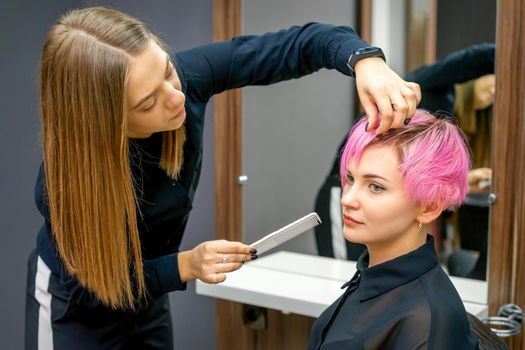 Female hairdresser styling short pink hair of the young white woman with hands and comb in a hair salon