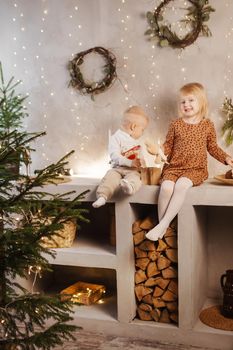 Little brother and sister play on Christmas eve in a beautiful house decorated for the New Year holidays. Children are playing with a Christmas gift. Scandinavian-style interior with live fir trees and a wooden staircase.