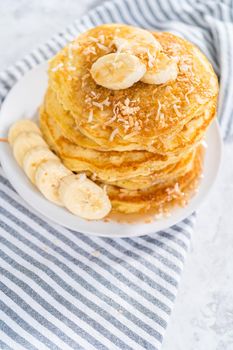 Stack of freshly baked coconut banana pancakes garnished with sliced bananas, toasted coconut, and maple syrup.