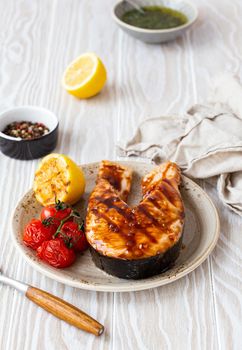 Grilled salmon steak glazed with teriyaki sauce, vegetables and lemon served on ceramic plate on rustic white wooden table from angle view, selective focus