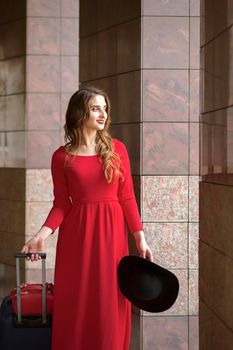 Beautiful young caucasian woman wearing red long dress with a suitcase standing in columns