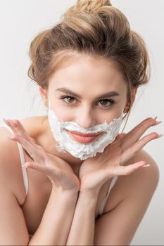 Portrait of a beautiful young smiling caucasian woman with shaving foam on face poses on white background