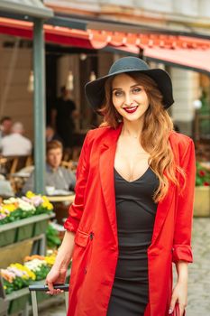 Smiling portrait of fashion young caucasian woman is posing in black hat and red jacket in the city street