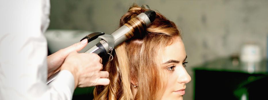 Hairdresser using curling tongs curls long brown hair on the young caucasian girl in a beauty salon