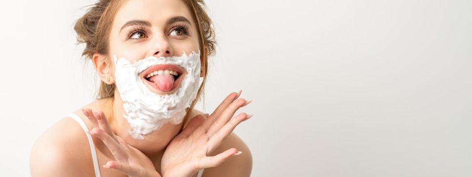 Portrait of a beautiful young smiling caucasian woman posing with shaving foam on the face stuck out her tongue on white background