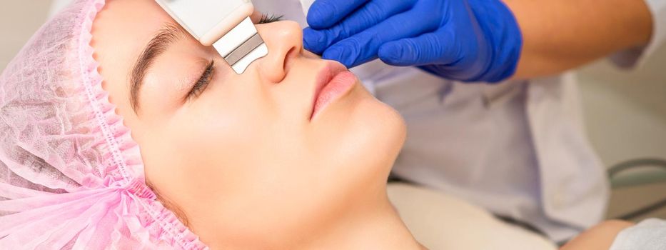 Closeup of beautiful young woman receiving ultrasound facial exfoliation and cavitation facial peeling with ultrasonic equipment in cosmetology office