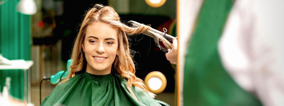 Hairdresser using curling tongs curls long brown hair on the young caucasian girl in a beauty salon