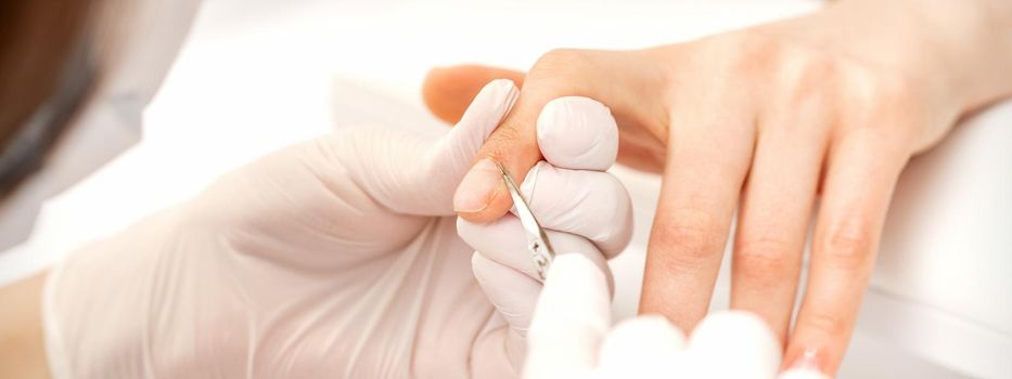 Close up of manicure master with manicure scissors removes cuticles of female nails at beauty salon