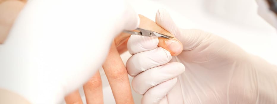 Close up of manicure master with manicure scissors removes cuticles of female nails at beauty salon