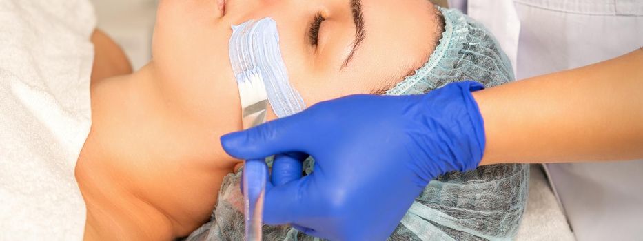 Beautician covering woman facial skin with moisturizing cleansing mask during skin care procedure in a spa beauty salon