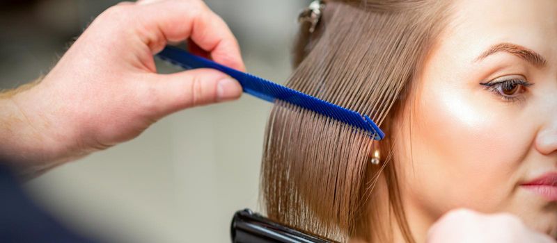 Hairdresser straightens female brown hair of medium length with a hair with an iron hair straightener and comb in a beauty salon