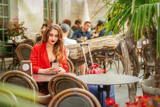 Tourist young caucasian white woman in a red jacket looking at smartphone sitting at the table in cafe outdoors