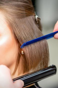 Hairdresser straightens female brown hair of medium length with a hair with an iron hair straightener and comb in a beauty salon