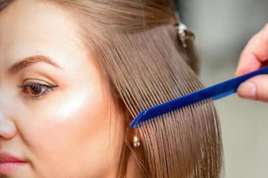 Hairdresser straightens female brown hair of medium length with a hair with an iron hair straightener and comb in a beauty salon