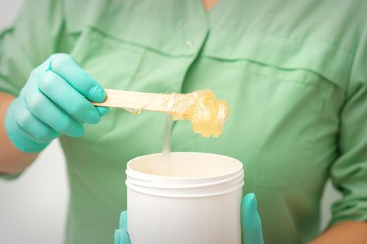 Liquid yellow sugar paste for depilation on a stick flows into the jar in hands of a beautician