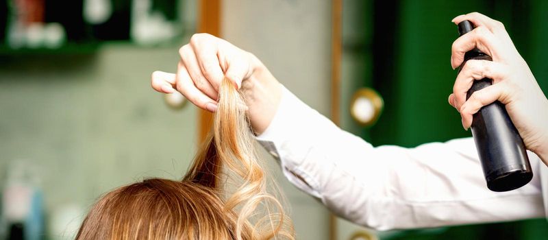 Female coiffeur fixing hairstyling of blonde woman with hairspray in a beauty salon