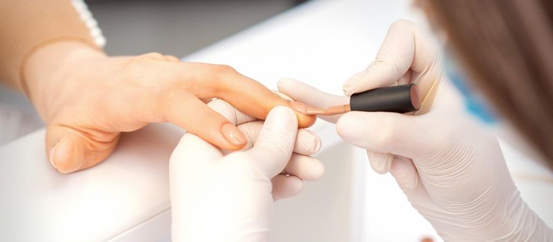 Hands of manicure master in white protective gloves apply polish to female fingernails