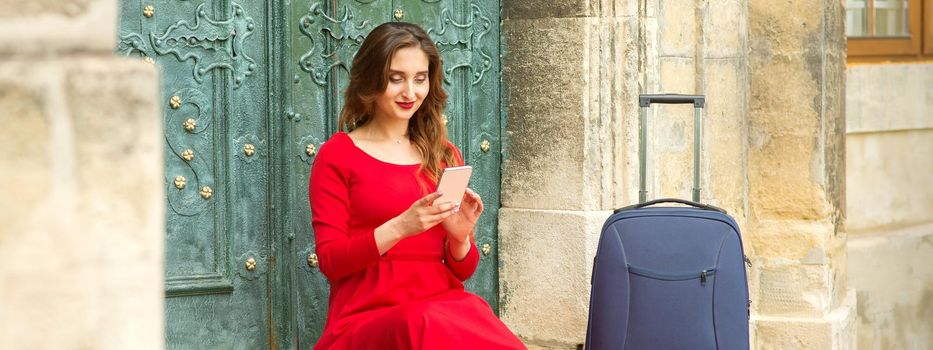 Beautiful young caucasian woman sitting on the stairs by the door with travel suitcase and smartphone wearing long red dress